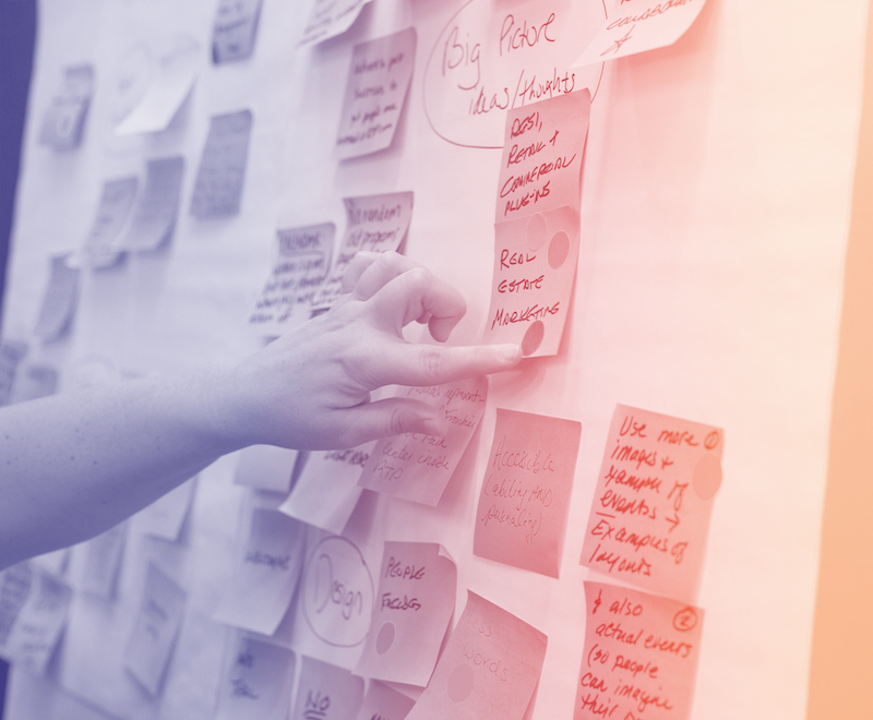 A person's hand pointing to a sticky note on a wall with other sticky notes.