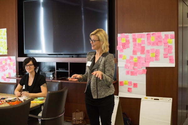 Christina is standing at the front of a conference room speaking with large sheets of paper with pink sticky notes behind her. Alisa is sitting at the head of the conference table watching Christina.