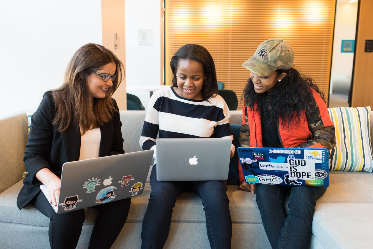 Three women using their laptops together on a couch. Two of them have programmer stickers on their laptops.