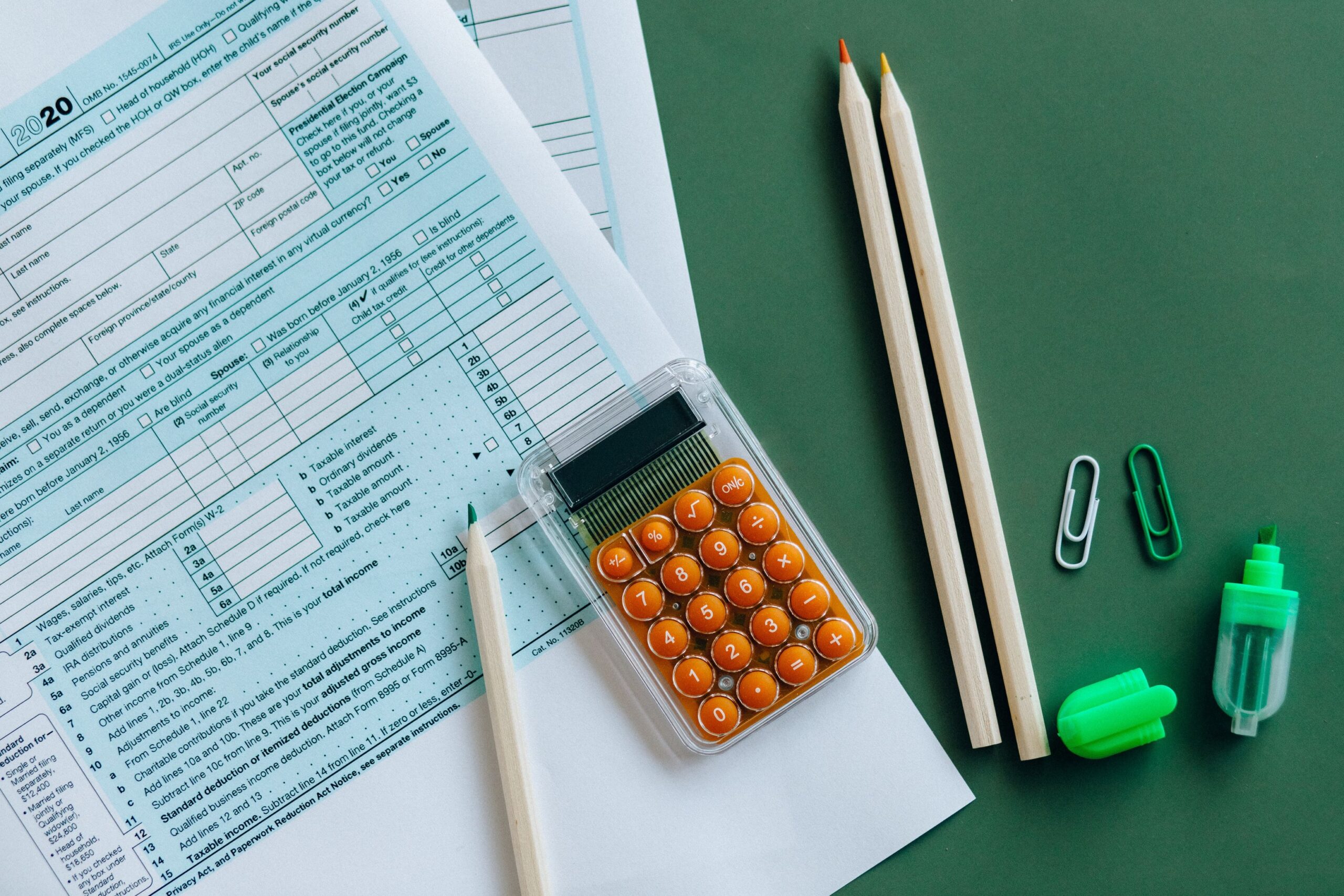 Photo of printed tax form on dark green surface. A handheld calculator with orange buttons, three wooden pencils, two paperclips, and a green highlighter are postioned on and beside the tax form. Photo by Nataliya Vaitkevich on Pexels.