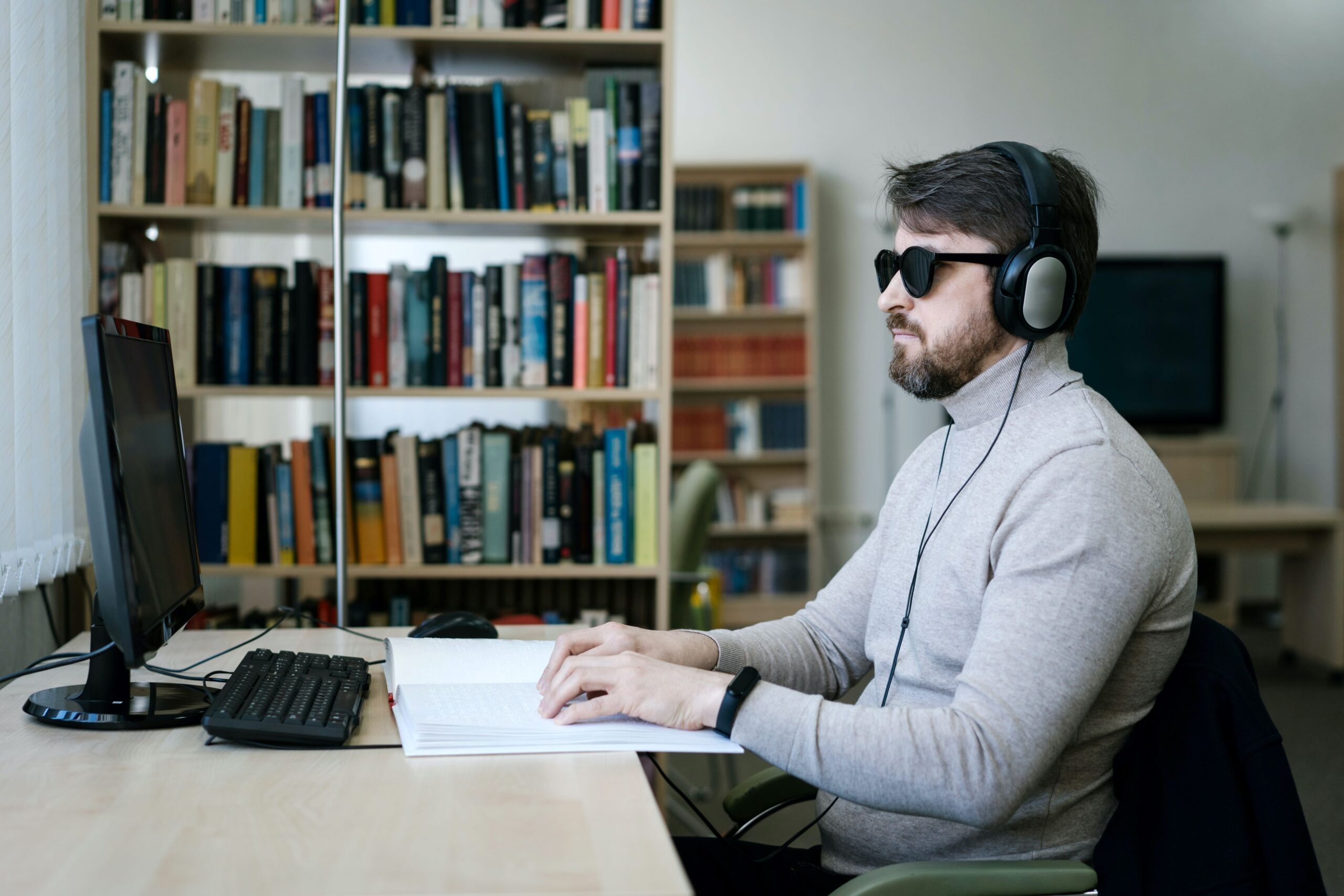 Photo of a blind person sitting at a desk in front of a computer. The man is wearing sunglasses and over-ear headphones. His hands are outstretched on a Braille book. There is a bookcase against the wall in the background. Photo by Nataliya Vaitkevich on Pexels.