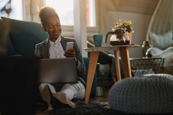 Black woman wearing a white button up and gray blazer is sitting comfortably at home while working on her laptop and checking her cell phone. A pot of coffee rests on a table next to her.